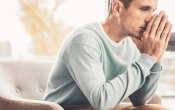 A man sitting on a gray chair, wearing a light blue sweater, with his hands pressed together against his face in a thoughtful or concerned manner. He looks towards the window, with soft natural light illuminating the scene.