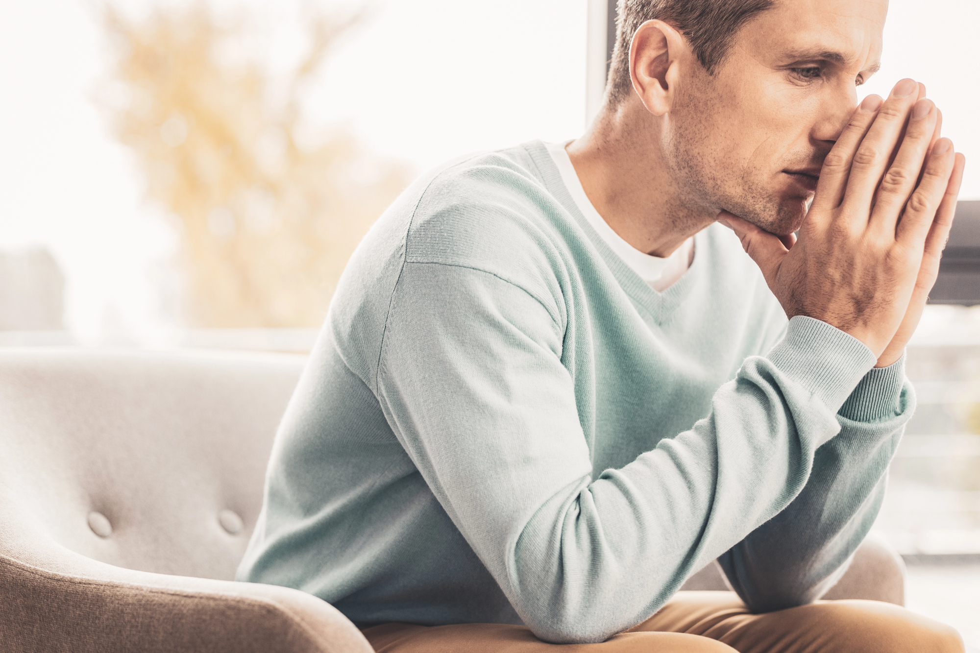 A man sitting on a gray chair, wearing a light blue sweater, with his hands pressed together against his face in a thoughtful or concerned manner. He looks towards the window, with soft natural light illuminating the scene.