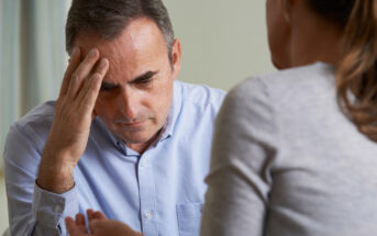 A man in a light blue shirt, appearing thoughtful or stressed, holds his forehead with one hand while facing a woman in a gray top. She gestures with her hands as if explaining something. They are sitting indoors, possibly engaged in a conversation.