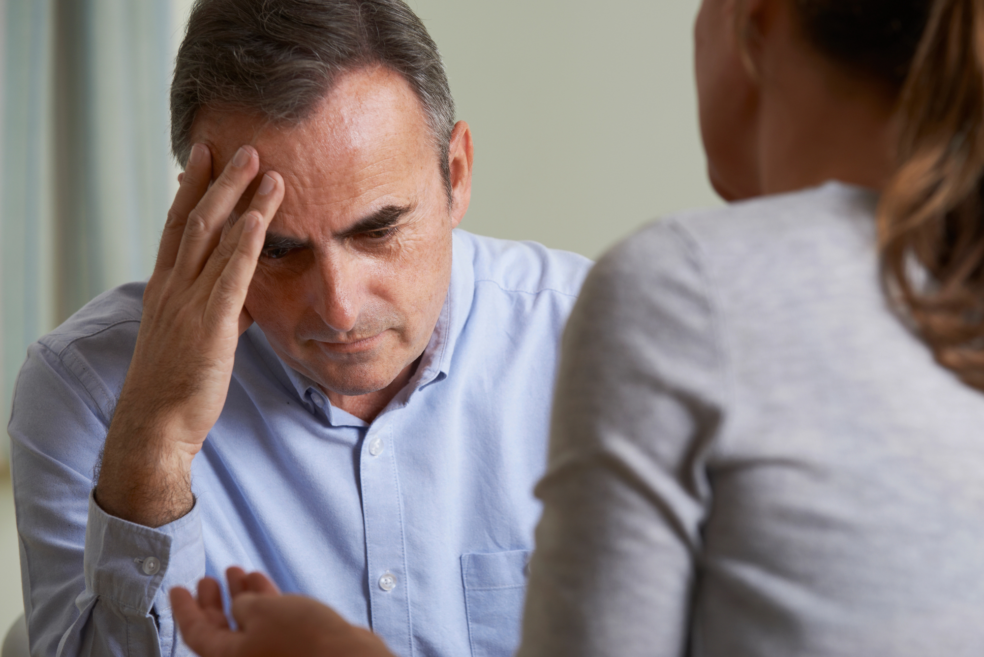 A man in a light blue shirt, appearing thoughtful or stressed, holds his forehead with one hand while facing a woman in a gray top. She gestures with her hands as if explaining something. They are sitting indoors, possibly engaged in a conversation.