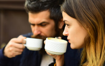 A man and a woman sit outdoors, both holding and sipping from white ceramic cups. The woman is in focus and has light brown hair, while the man in the background has a beard. They appear to be enjoying a warm beverage.