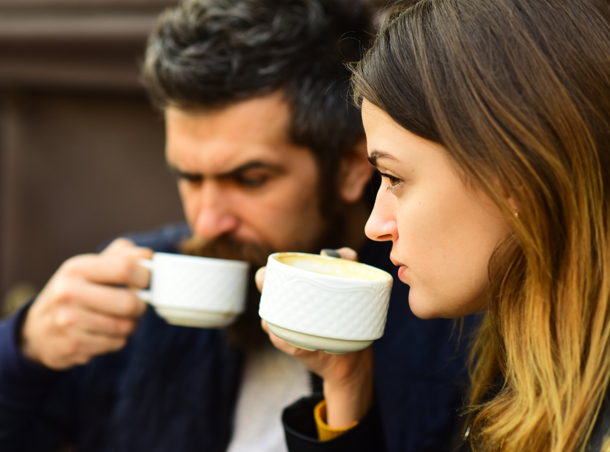 A man and a woman sit outdoors, both holding and sipping from white ceramic cups. The woman is in focus and has light brown hair, while the man in the background has a beard. They appear to be enjoying a warm beverage.