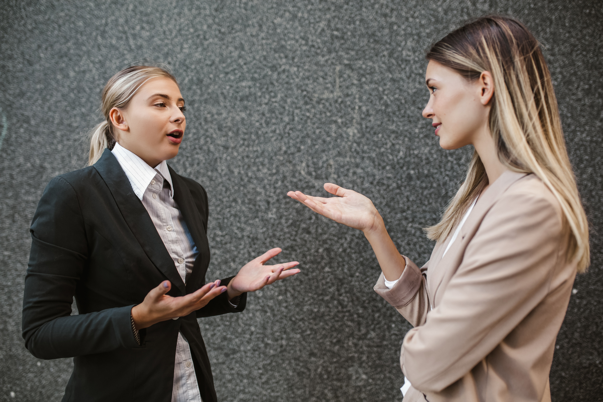 Two women in business attire are engaged in a conversation in front of a gray textured wall. One gestures with open hands, while the other listens attentively, hand raised. Both appear focused and professional.