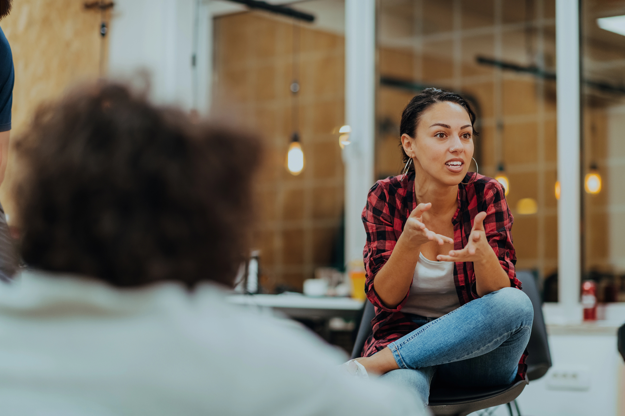 A woman in a red and black plaid shirt and jeans sits on a chair, engaged in conversation, gesturing with her hands. She is in a modern office space with a blurred person in the foreground.