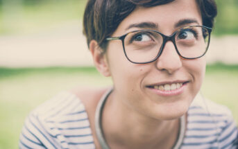 A person with short brown hair and glasses is smiling while looking to the side. They are wearing a striped shirt and are outdoors with a blurred green background.