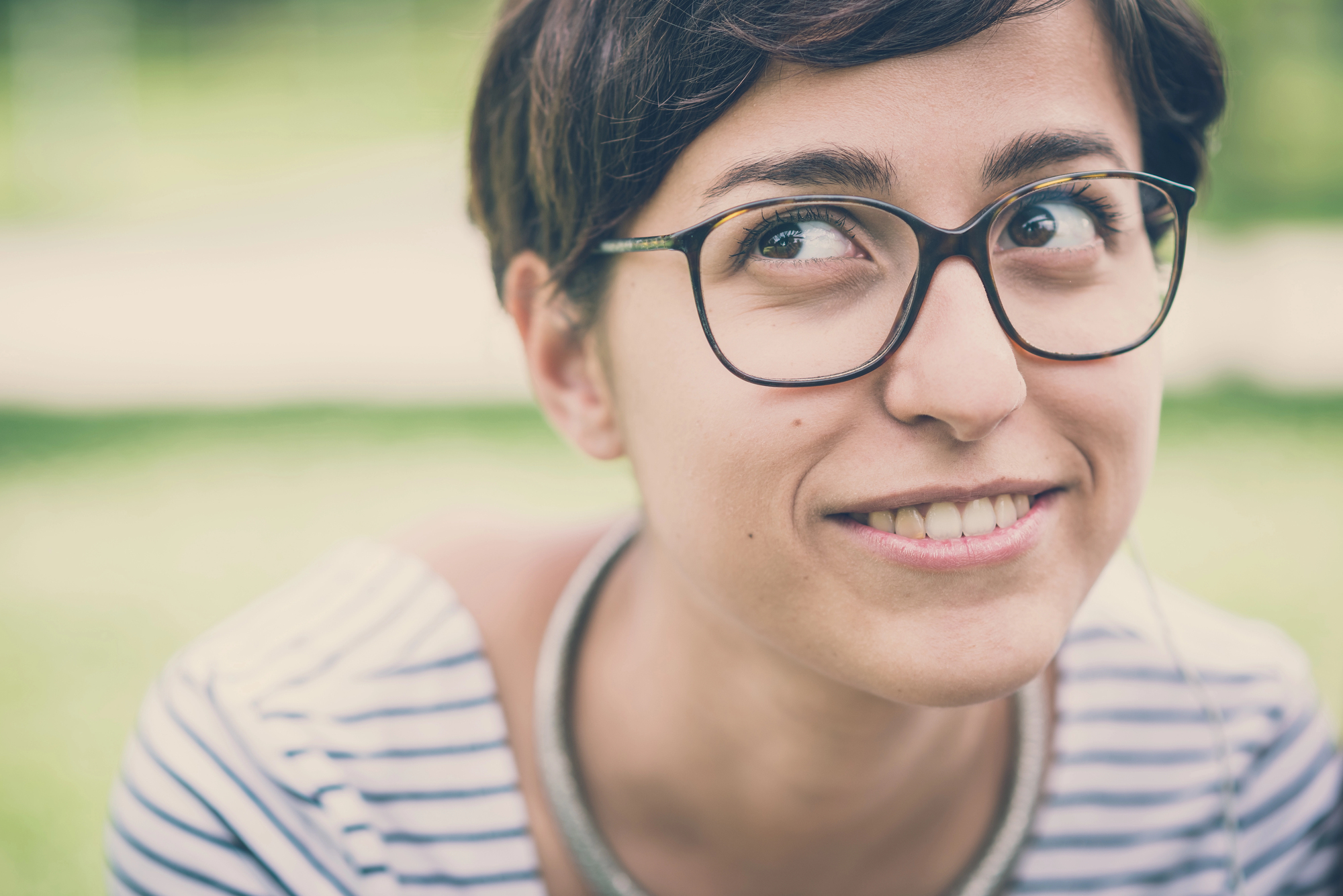 A person with short brown hair and glasses is smiling while looking to the side. They are wearing a striped shirt and are outdoors with a blurred green background.