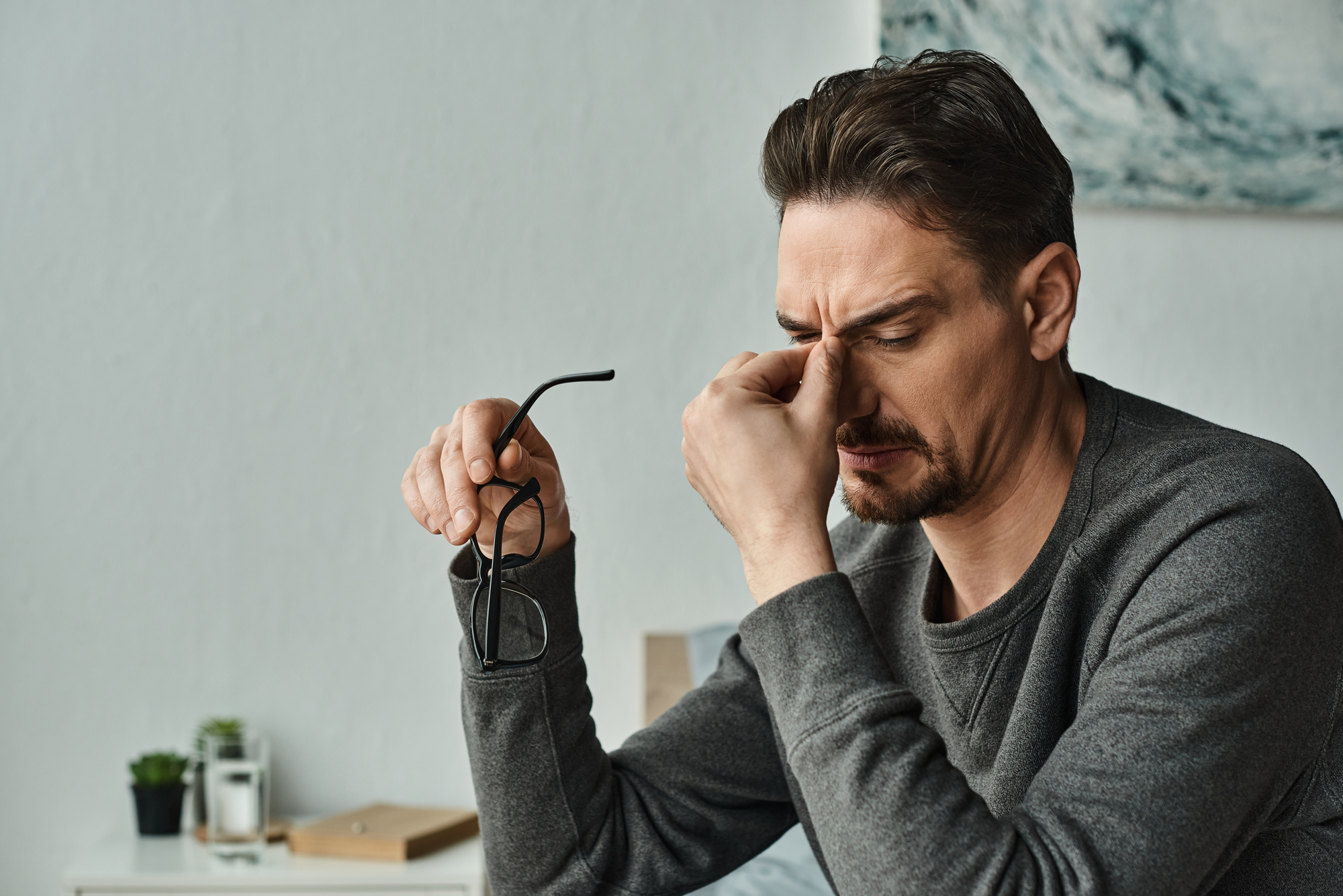 A man wearing a gray sweater is sitting indoors, holding his glasses in one hand and rubbing his eyes with the other, appearing tired or stressed. Behind him are a small potted plant and some books on the table.