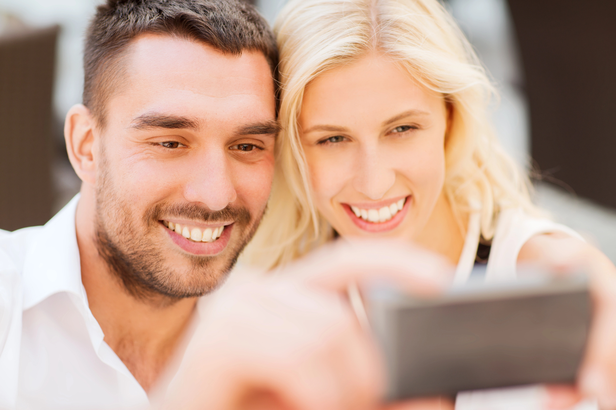 A man and woman smiling as they take a selfie together. The man has short dark hair and a beard, wearing a white shirt. The woman has long blonde hair and is also smiling brightly. They appear to be outdoors.