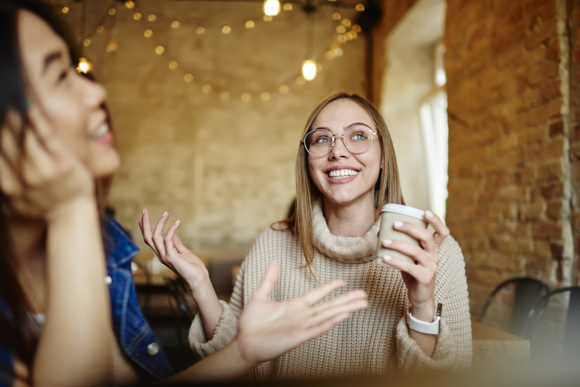 Two people are sitting at a cozy cafe, engaged in a lively conversation. One is holding a coffee cup and smiling, while the other gestures expressively. Warm lighting and exposed brick walls create a relaxed atmosphere.