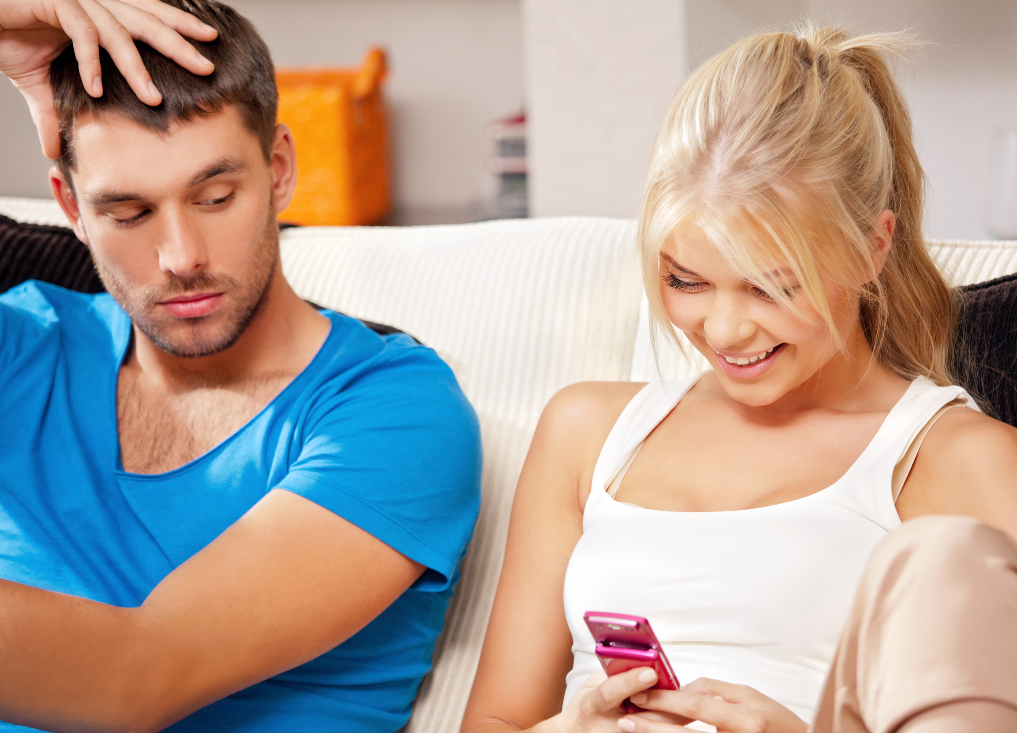 A woman with blonde hair, wearing a white tank top, is sitting on a couch and smiling at her pink phone. Next to her, a man with short dark hair in a blue shirt looks over at her with a curious expression.