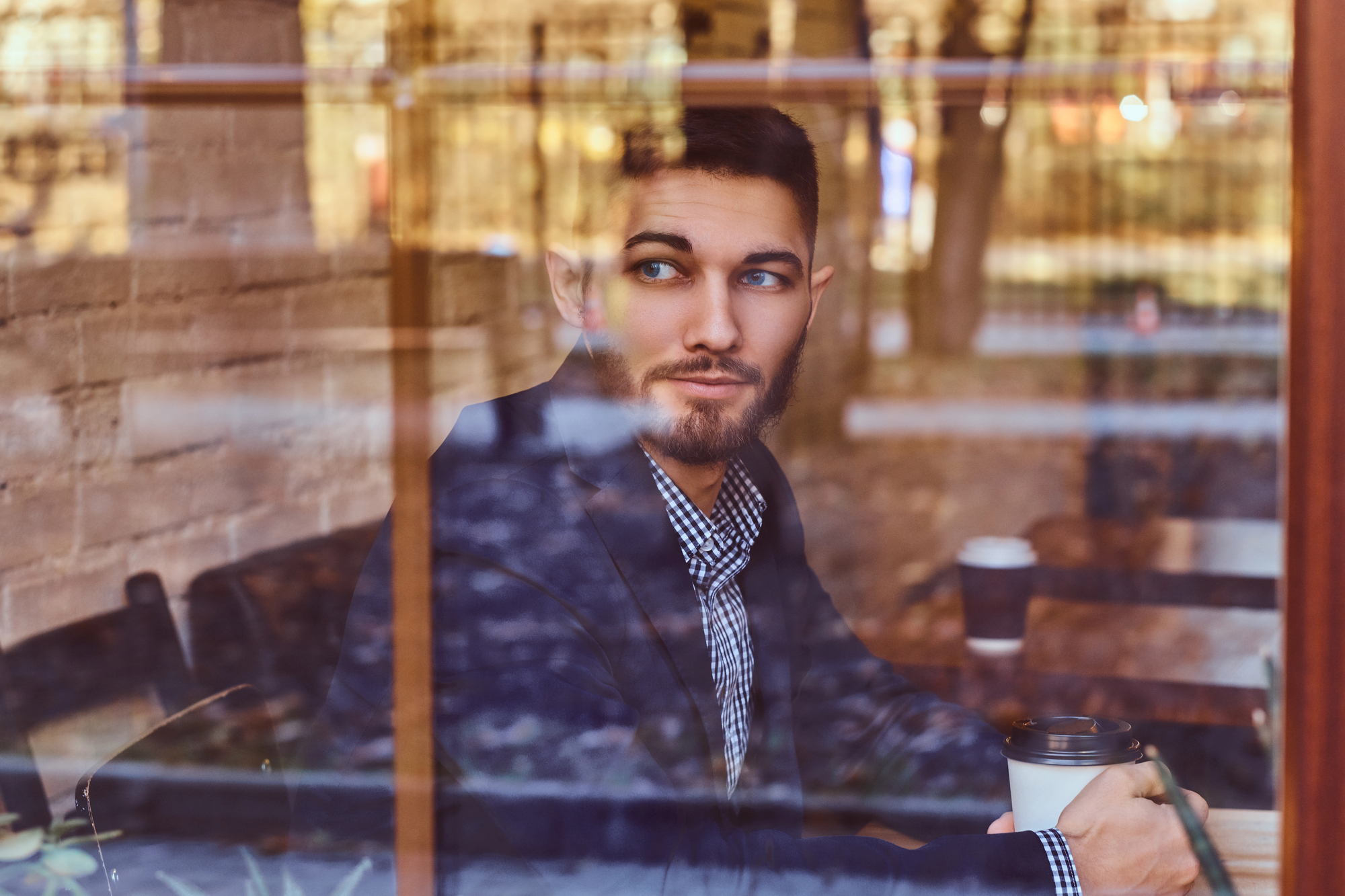 A man with a beard sits in a cafe holding a coffee cup, gazing thoughtfully out the window. The scene is reflected on the glass, showing blurred outdoor elements and warm, ambient lighting inside.