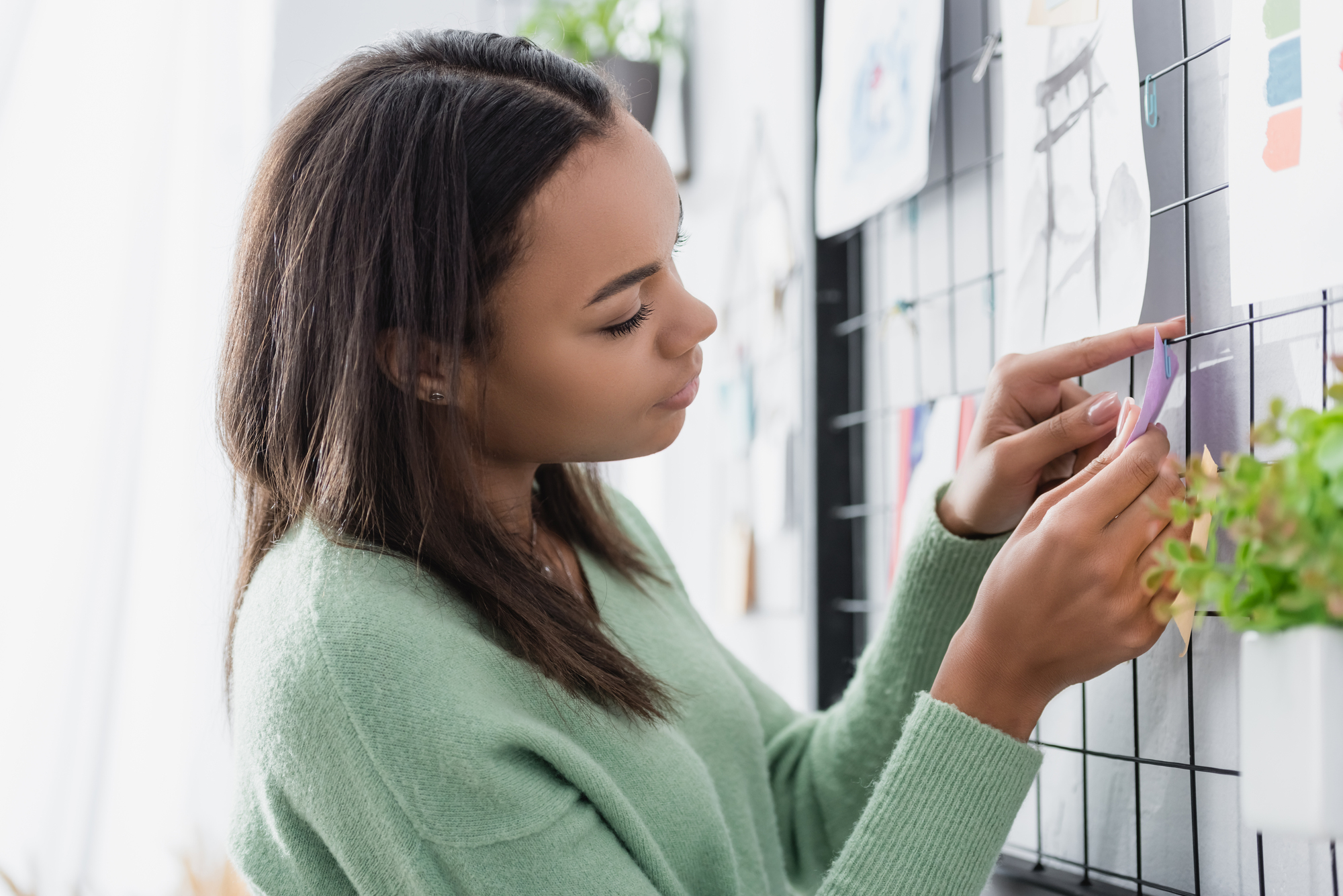 A woman wearing a green sweater is adding a note to a grid-like wall with various papers and images. She appears focused and is in a bright room with plants in the background.