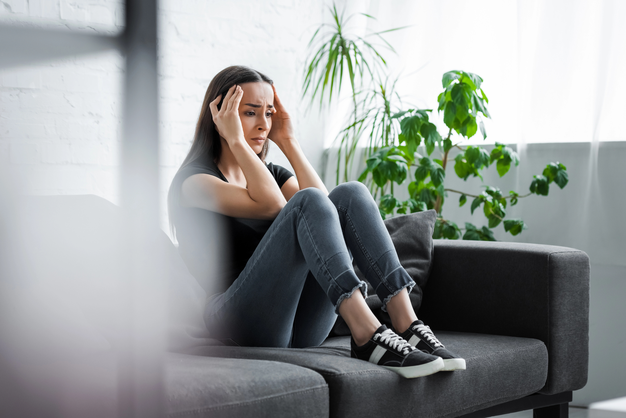 A woman sits on a sofa with her knees up, holding her head in her hands, looking distressed. She wears a black shirt, jeans, and sneakers. The room is bright with green plants in the background.