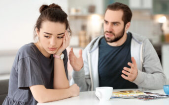 A woman with a concerned expression sits at a table with her hand on her face, while a man beside her gestures as if explaining something. There are a magazine and a white cup on the table in a kitchen setting.