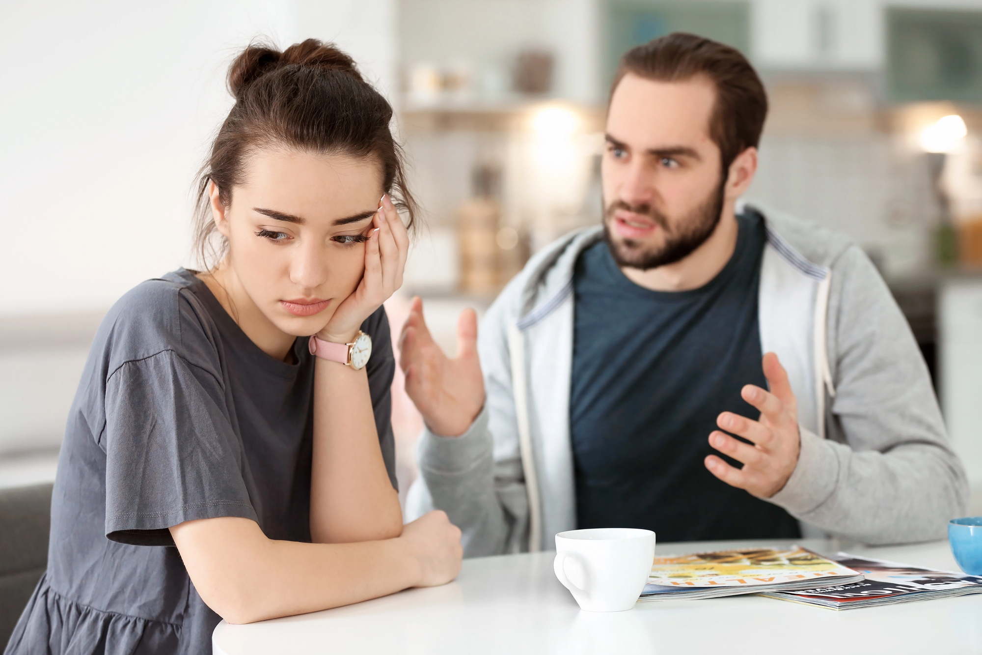 A woman with a concerned expression sits at a table with her hand on her face, while a man beside her gestures as if explaining something. There are a magazine and a white cup on the table in a kitchen setting.