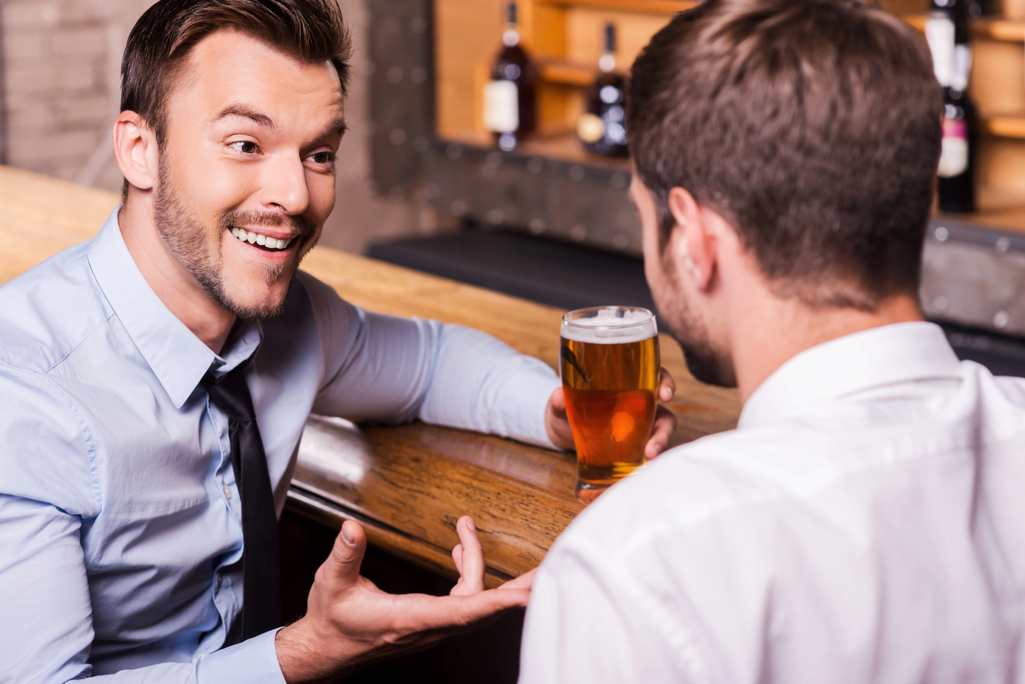 Two men in dress shirts sit at a bar counter. One faces the camera with a smile and gesturing hands, while the other, seen from behind, holds a glass of beer. Bottles are visible on the shelf in the background.