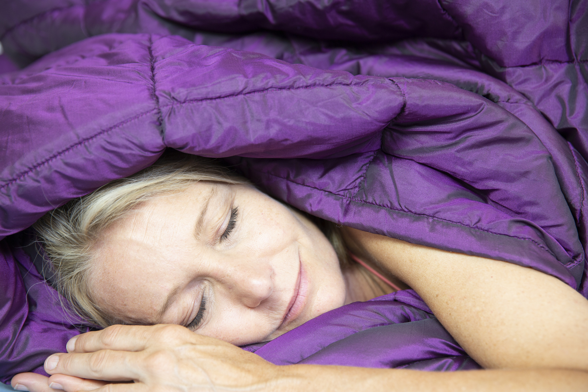 A person with light hair is peacefully sleeping under a fluffy, purple blanket, with a calm expression and eyes closed. Their hand rests near their face, and the blanket appears warm and cozy.