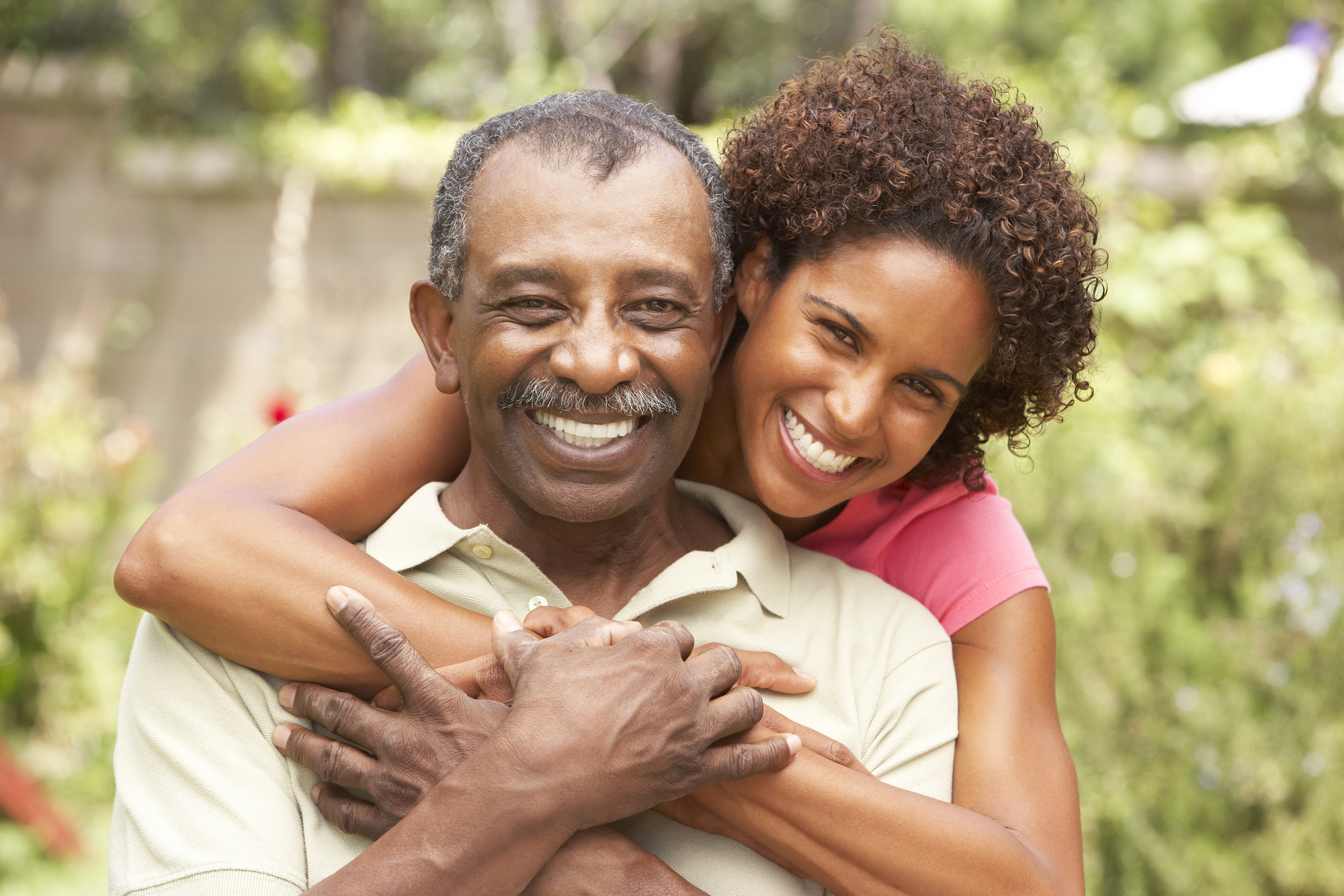 A woman embraces an older man from behind, both smiling warmly. They are outdoors with lush greenery in the background, creating a joyful and vibrant atmosphere.