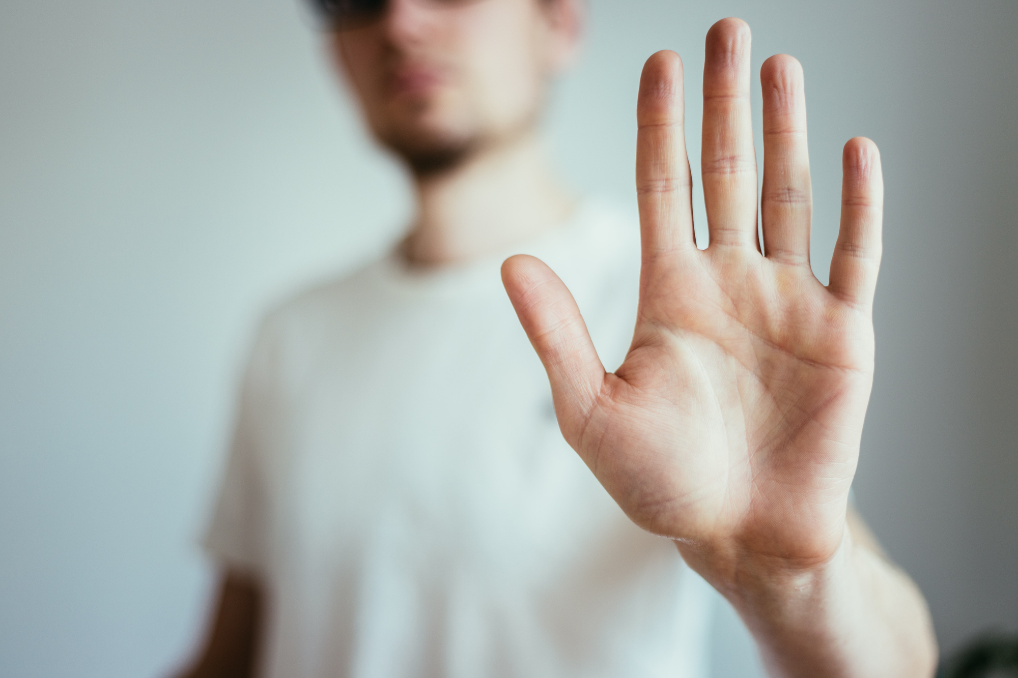 A man in a white t-shirt extends his hand toward the camera, palm facing forward, in a stop gesture. The background is softly blurred, emphasizing the hand's detail.