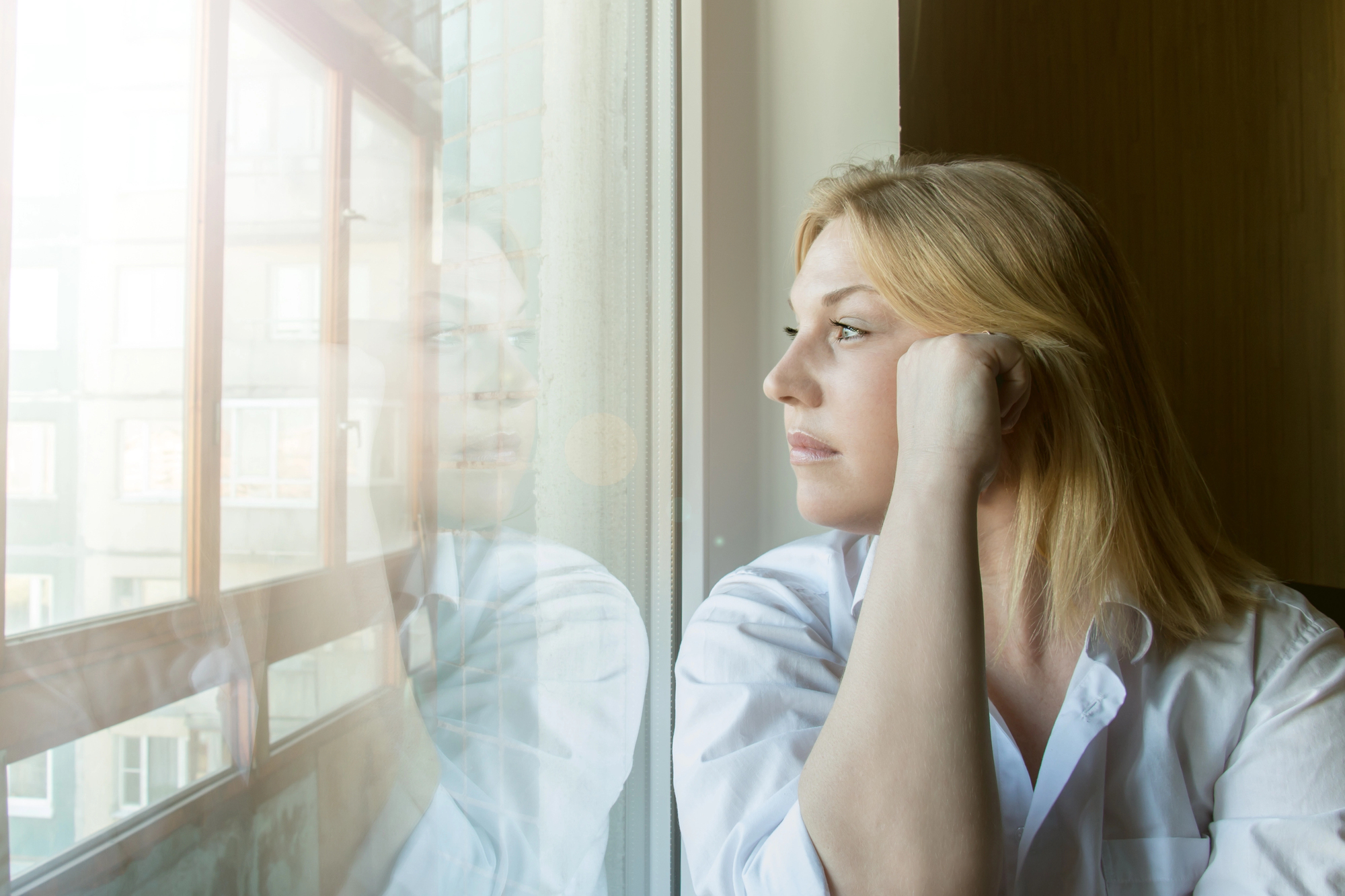 A woman with long blond hair wearing a white shirt sits by a window, resting her head on her hand. She gazes thoughtfully outside, with her reflection visible in the glass. A cityscape is faintly visible through the window.