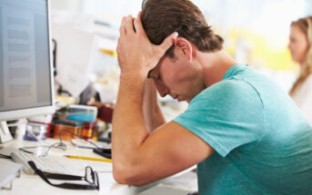 A man in a teal shirt sits at a desk with his head in his hands, appearing stressed. A computer monitor displays documents, and a pair of glasses rests on the table. A woman works in the background in an office setting.