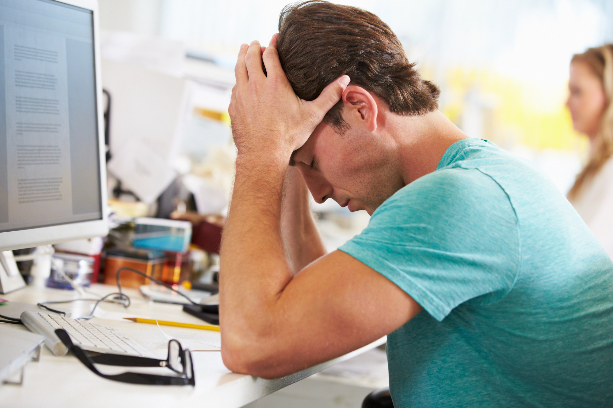 A man in a teal shirt sits at a desk with his head in his hands, appearing stressed. A computer monitor displays documents, and a pair of glasses rests on the table. A woman works in the background in an office setting.