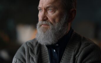 Elderly man with a gray beard and serious expression gazes to the side. He is wearing a dark shirt and a knitted cardigan. The background is softly blurred.