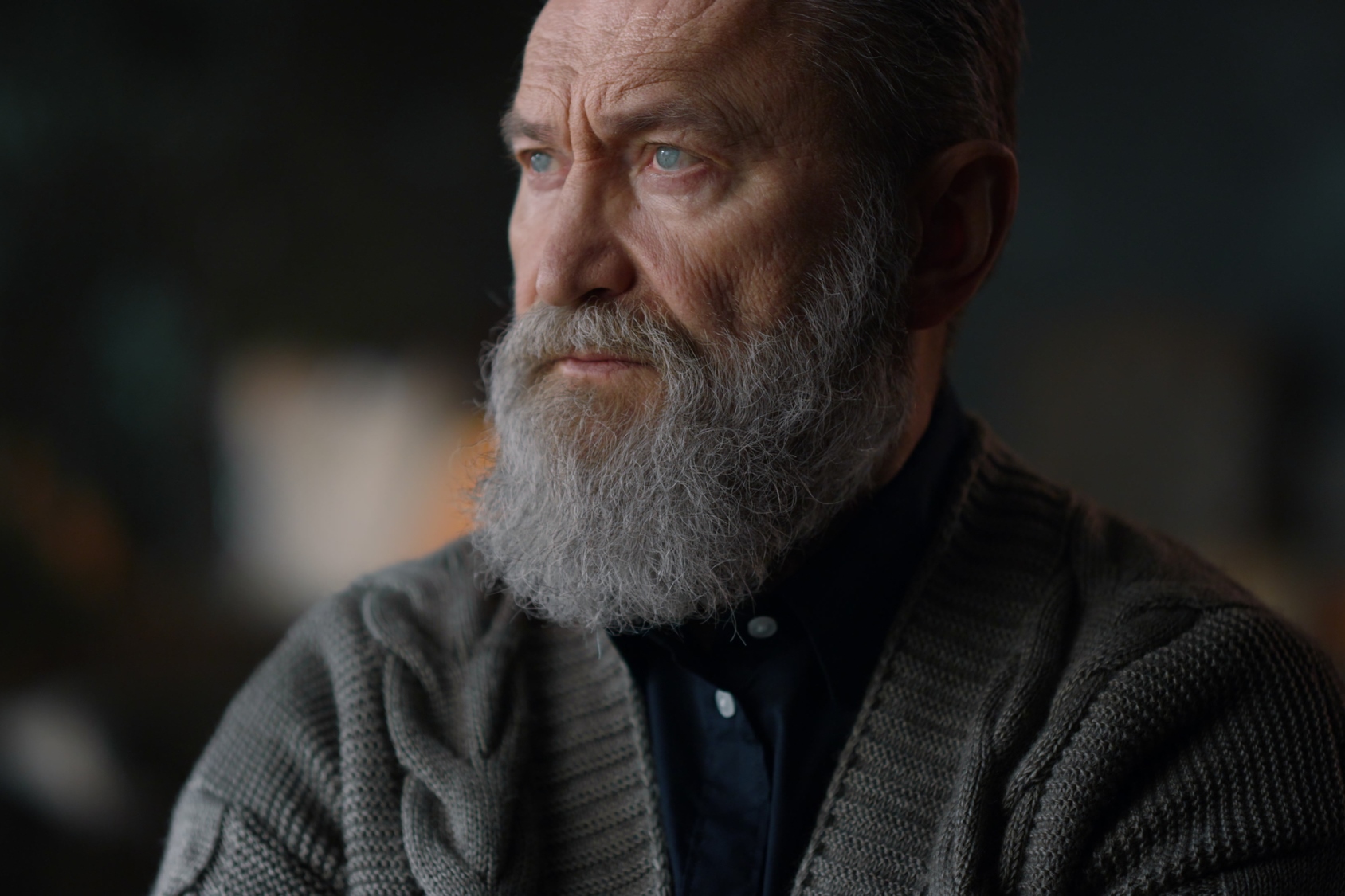 Elderly man with a gray beard and serious expression gazes to the side. He is wearing a dark shirt and a knitted cardigan. The background is softly blurred.