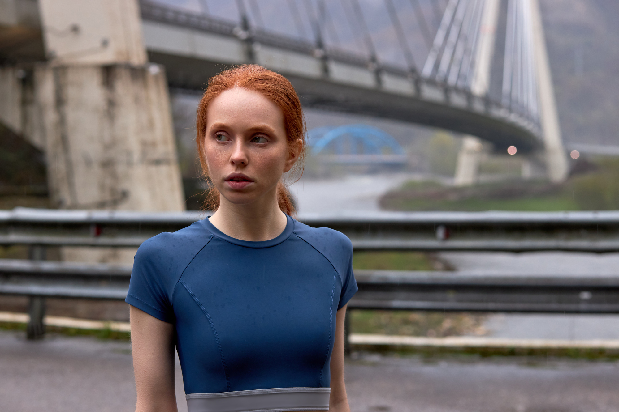 A woman with red hair wearing a blue athletic outfit stands near a river with a large bridge in the background. The scene is overcast, and the bridge structure is prominent in the image.