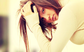 A woman with long brown hair, wearing a beige sweater, rests her head in her hand, looking pensively at the camera. The background is softly blurred, creating a serene and contemplative atmosphere.