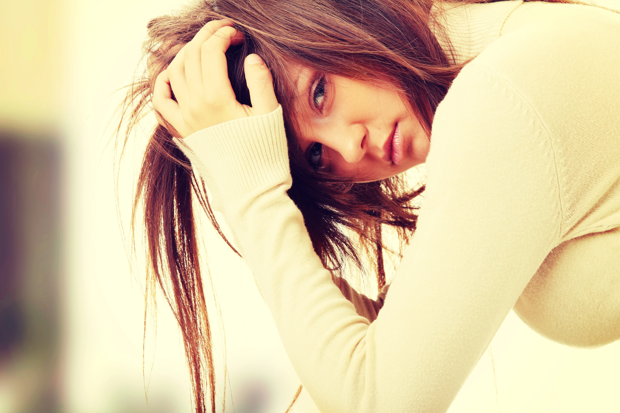 A woman with long brown hair, wearing a beige sweater, rests her head in her hand, looking pensively at the camera. The background is softly blurred, creating a serene and contemplative atmosphere.