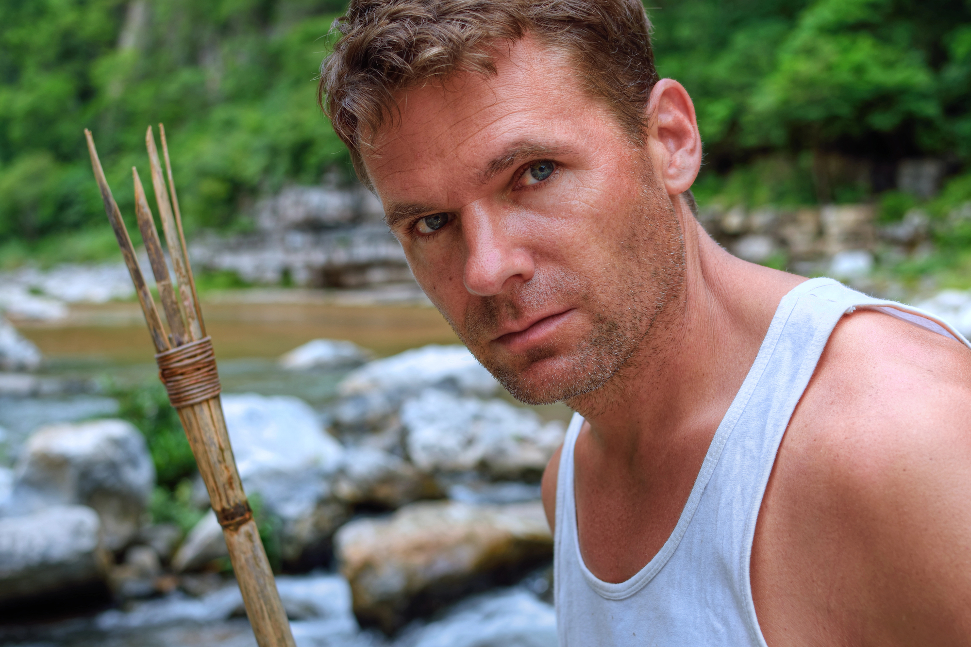 A man in a white tank top holds a wooden spear, standing near rocks and a river. He is outdoors, surrounded by lush greenery. His expression is focused and intense.