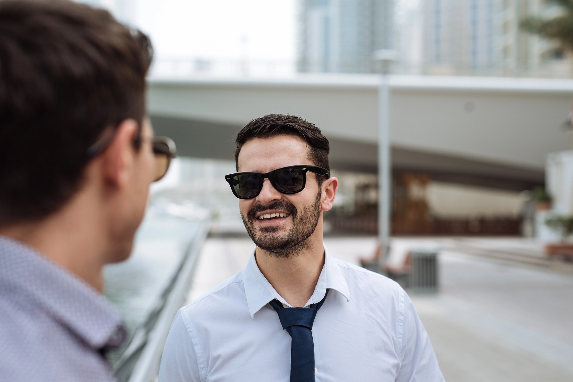 Two men outdoors in an urban setting. The man in the foreground is wearing sunglasses, a white shirt, and a blue tie, smiling at the other man, who is out of focus. A bridge and buildings are visible in the background.