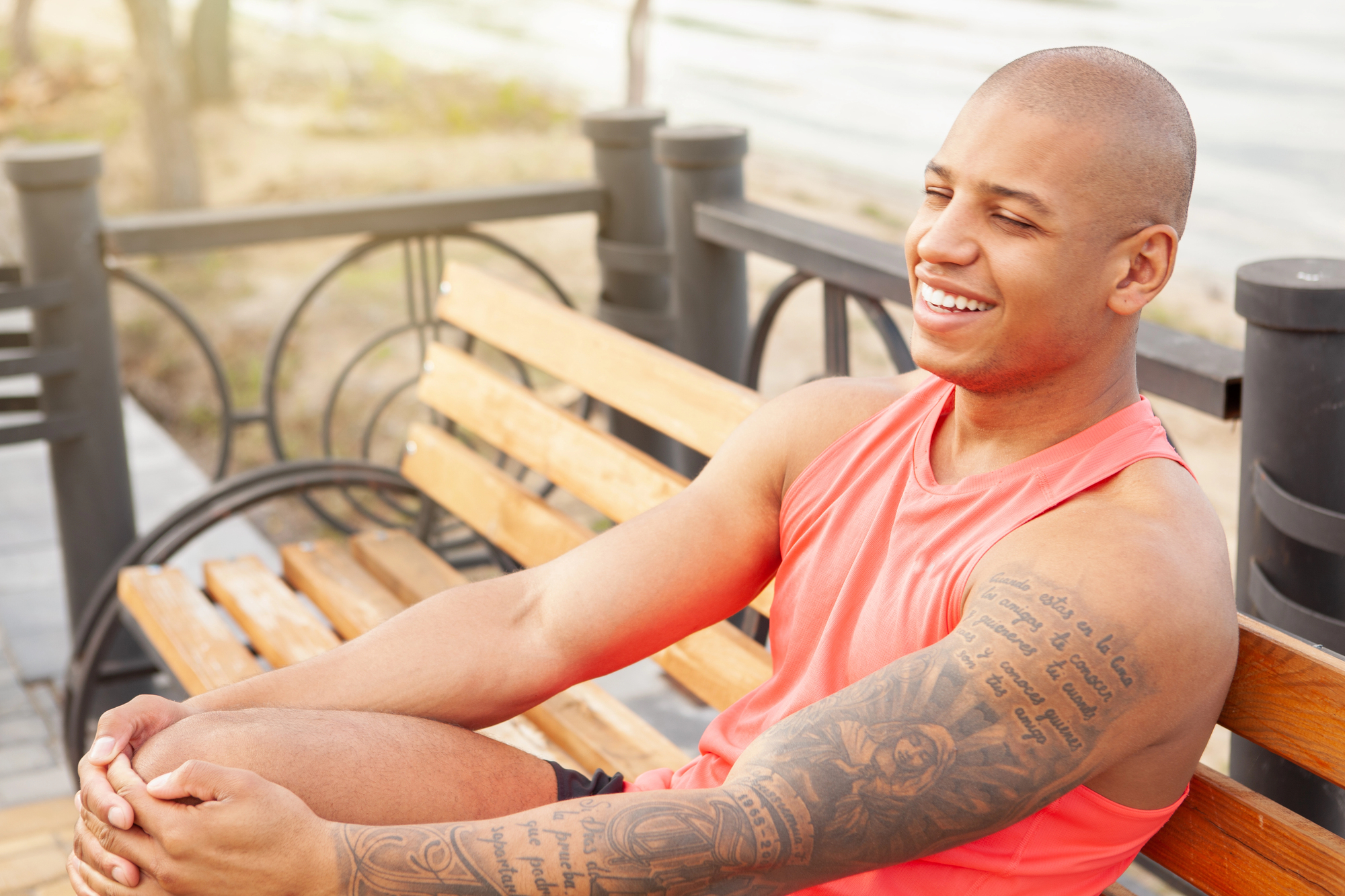 A smiling man with a tattooed arm sits on a wooden bench outdoors in a park. He is wearing a sleeveless orange shirt and black shorts. The background shows a sunny day with trees and a pathway.