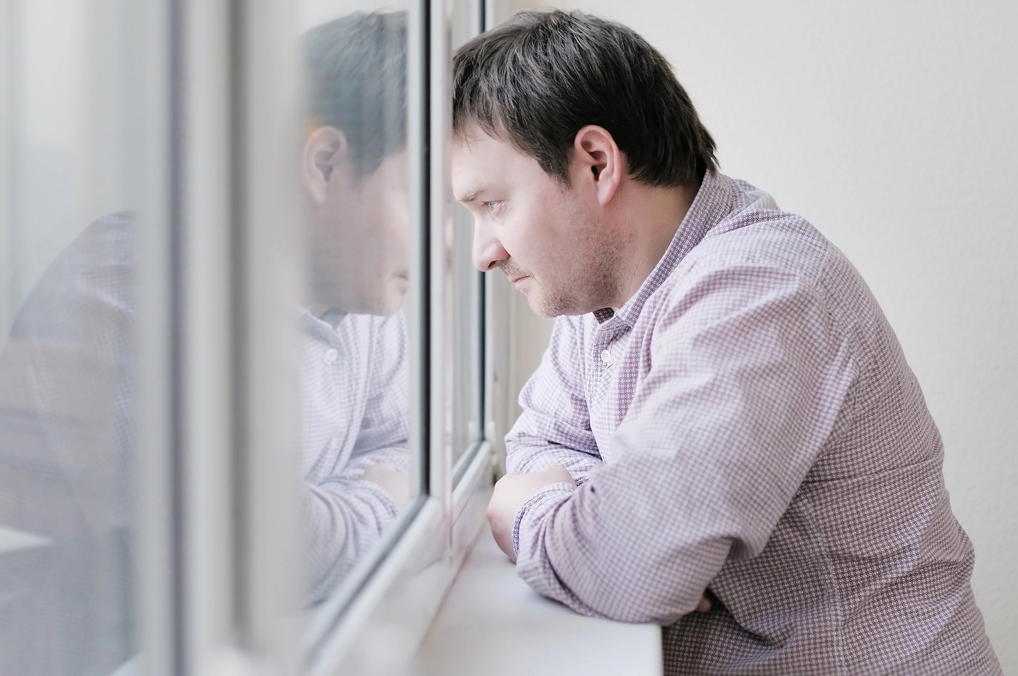 A man in a checkered shirt leans on a windowsill, looking contemplative. His reflection is visible in the glass. The room is softly lit, creating a calm and introspective atmosphere.