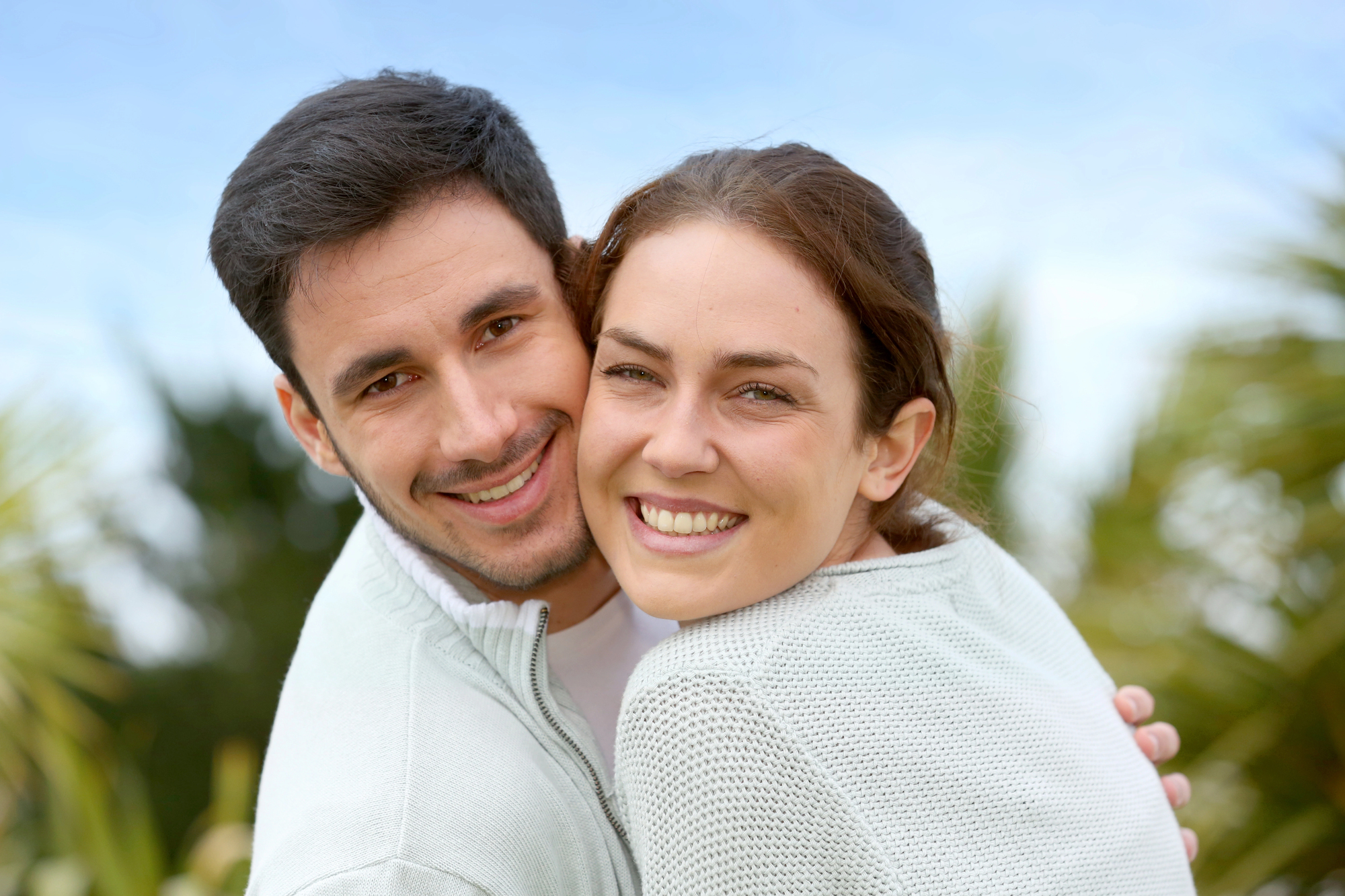 A smiling couple embraces outdoors. They are wearing light-colored sweaters and appear happy. The background shows a blurred view of greenery and a clear blue sky.