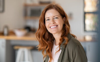 A woman with long red hair smiles warmly while sitting in a bright kitchen. She is wearing a light-colored shirt and a green jacket. The background shows blurred kitchen shelves and a counter.