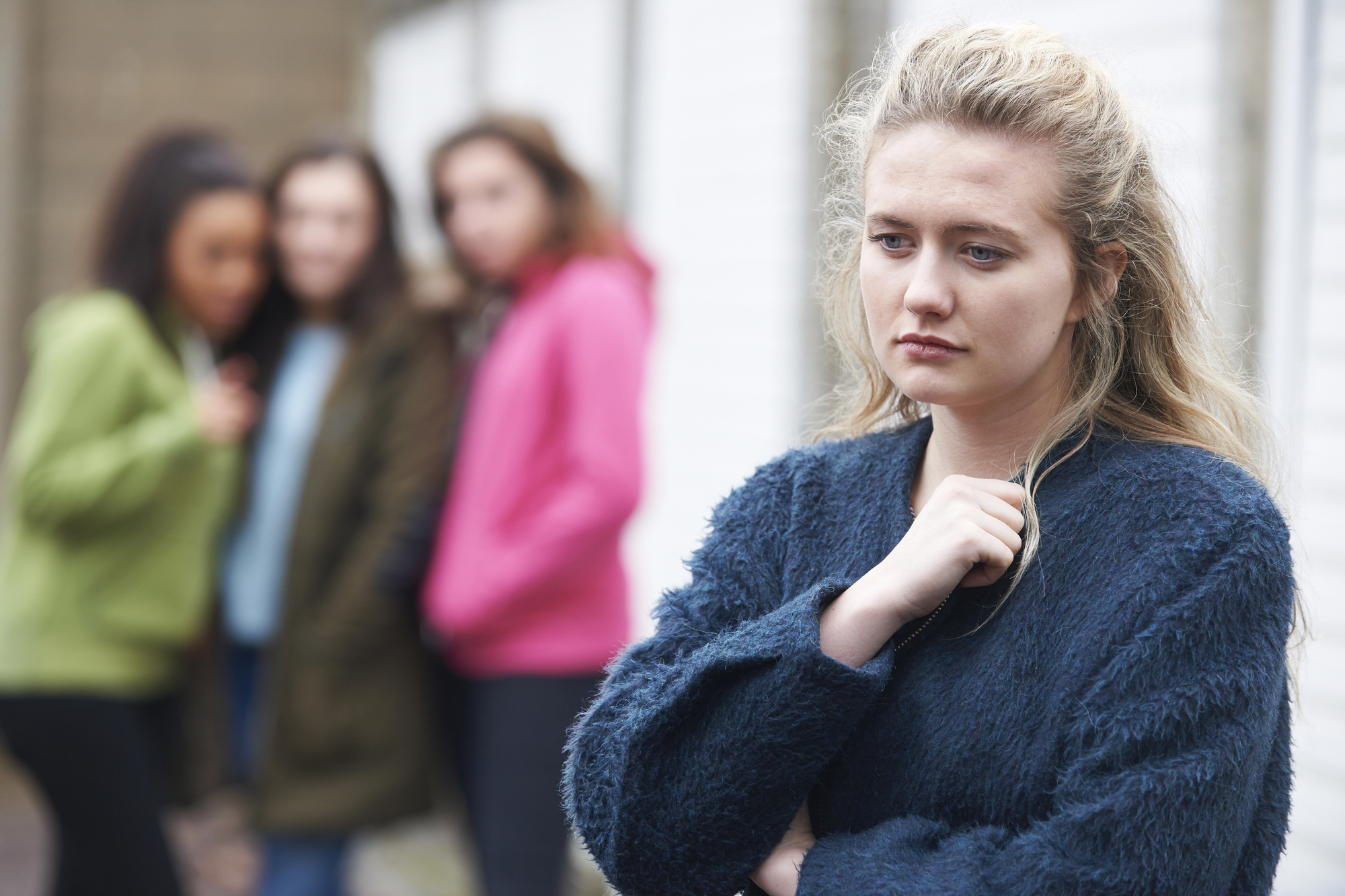 A young woman in a blue sweater looks upset and thoughtful in the foreground. In the background, three other women are standing together, slightly out of focus, suggesting they may be talking about her.