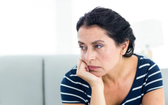 A woman with dark hair is seated, resting her chin on her hand. She appears thoughtful and is wearing a navy blue and white striped shirt. The background is softly blurred, featuring a light-colored interior.