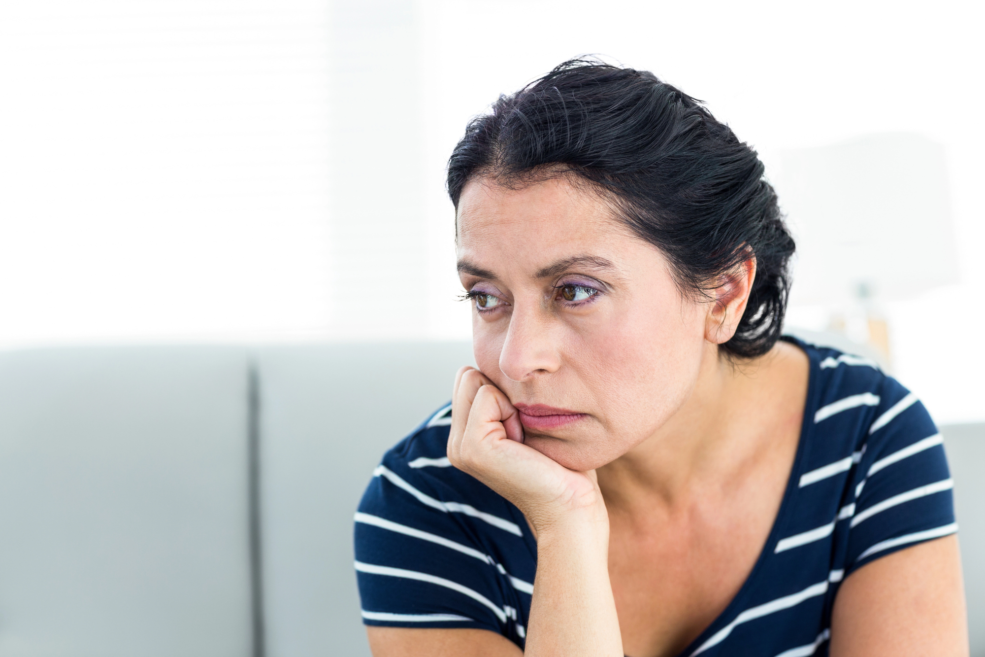 A woman with dark hair is seated, resting her chin on her hand. She appears thoughtful and is wearing a navy blue and white striped shirt. The background is softly blurred, featuring a light-colored interior.