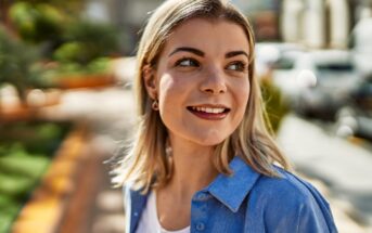 A woman with shoulder-length blonde hair smiles and looks to the side while standing outdoors. She is wearing a blue shirt over a white top. The background shows a blurred street scene with greenery and buildings.