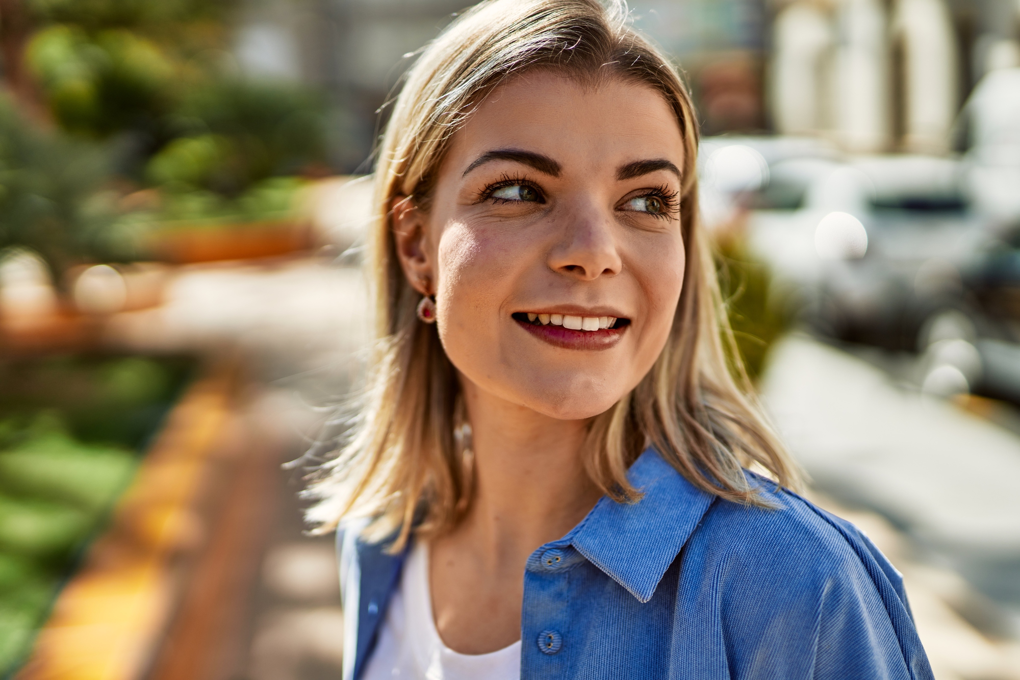 A woman with shoulder-length blonde hair smiles and looks to the side while standing outdoors. She is wearing a blue shirt over a white top. The background shows a blurred street scene with greenery and buildings.