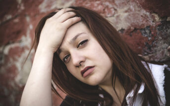 A woman with long brown hair and a nose ring looks pensively at the camera, resting her hand on her head. She stands against a textured, red brick wall, wearing a white-collared shirt.