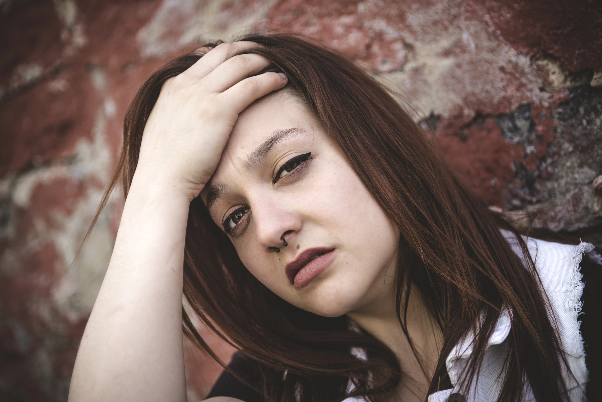 A woman with long brown hair and a nose ring looks pensively at the camera, resting her hand on her head. She stands against a textured, red brick wall, wearing a white-collared shirt.