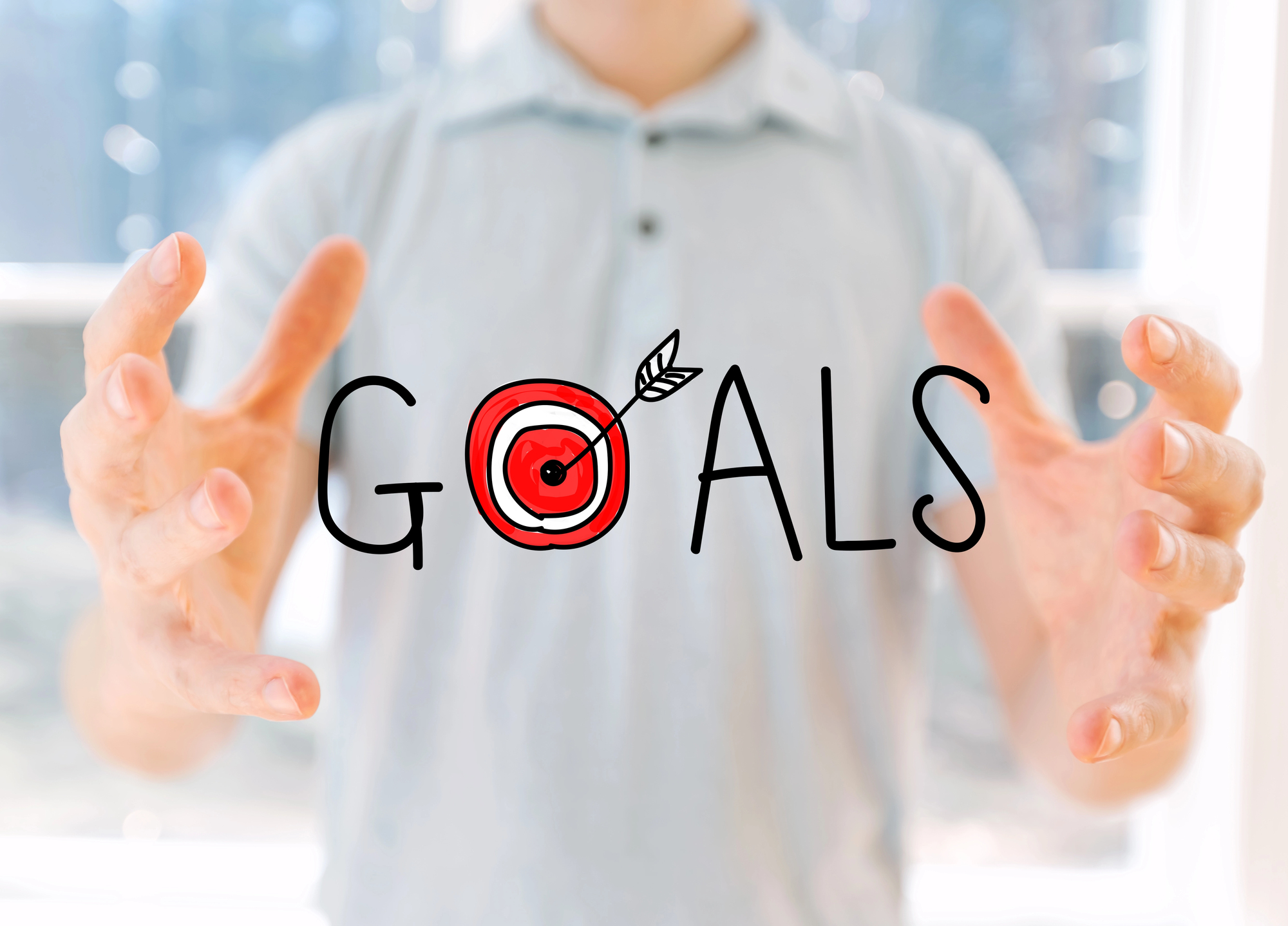 A person in a light shirt holds their hands open around the word "GOALS," written in black letters, with an illustration of a red target and an arrow replacing the letter "O." The background is bright and blurred.