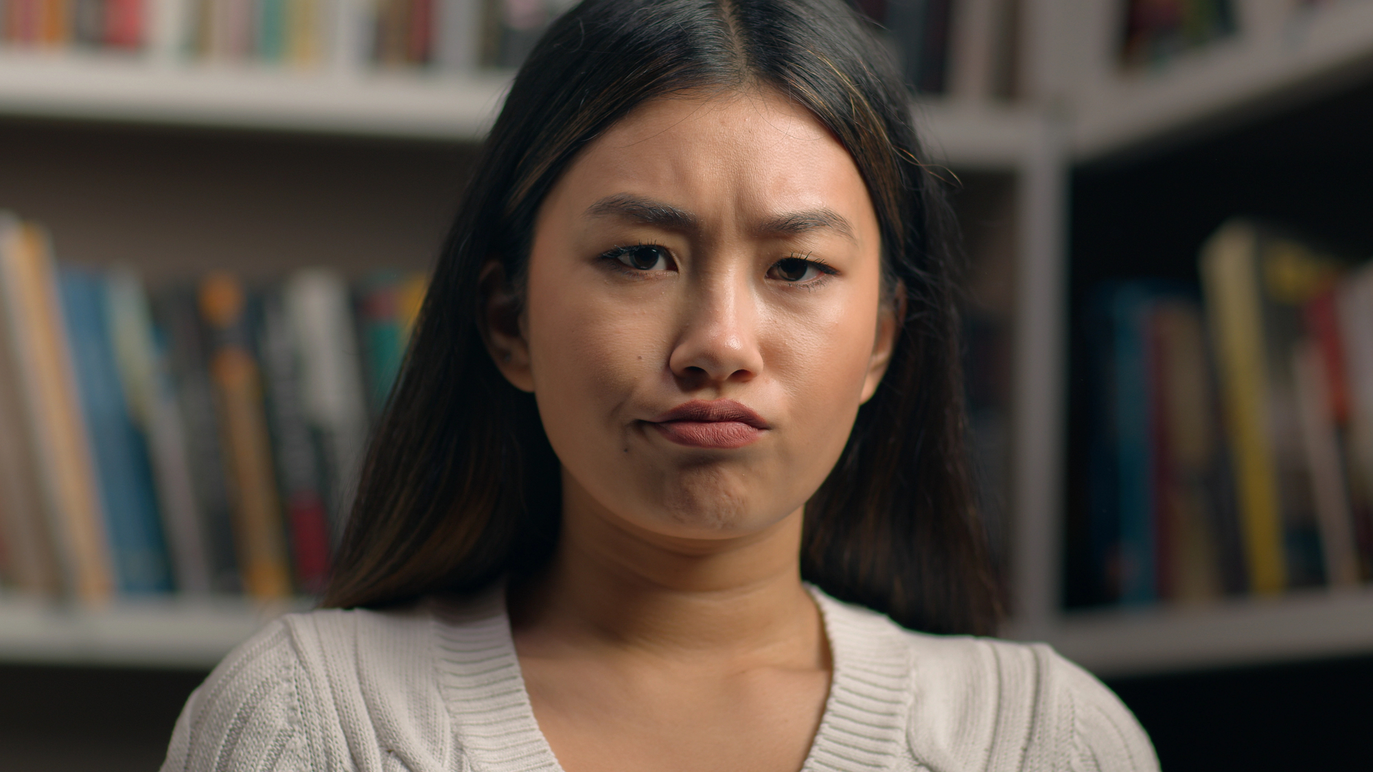 A woman with long dark hair and a white sweater is furrowing her brows and pursing her lips. She is sitting in front of a bookshelf filled with books.