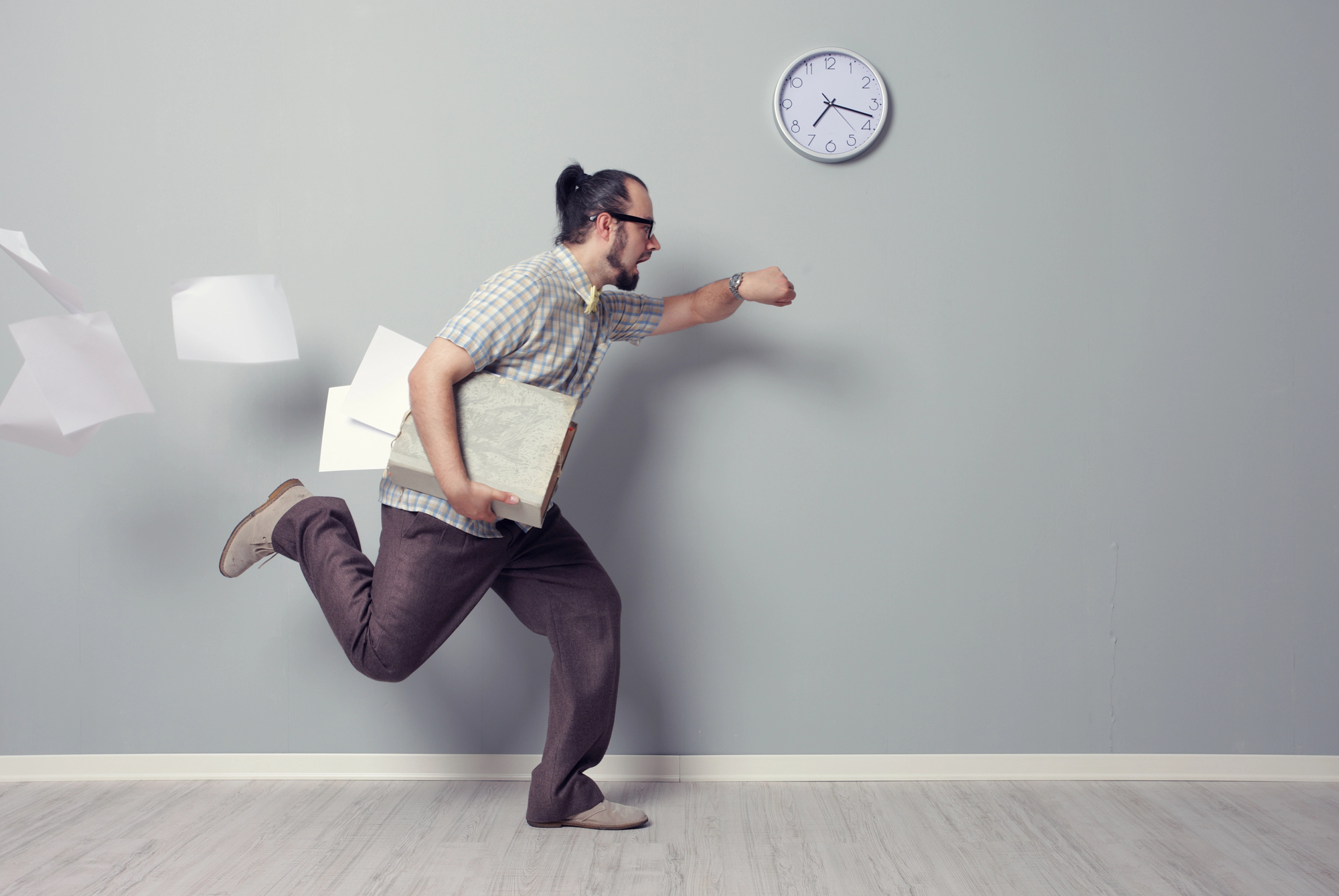 A man with a ponytail, wearing glasses and casual clothes, runs hurriedly while holding a folder and some papers, with a few sheets flying behind him. A clock on the wall shows 10:10. The setting is a minimalistic room with a light-colored floor.