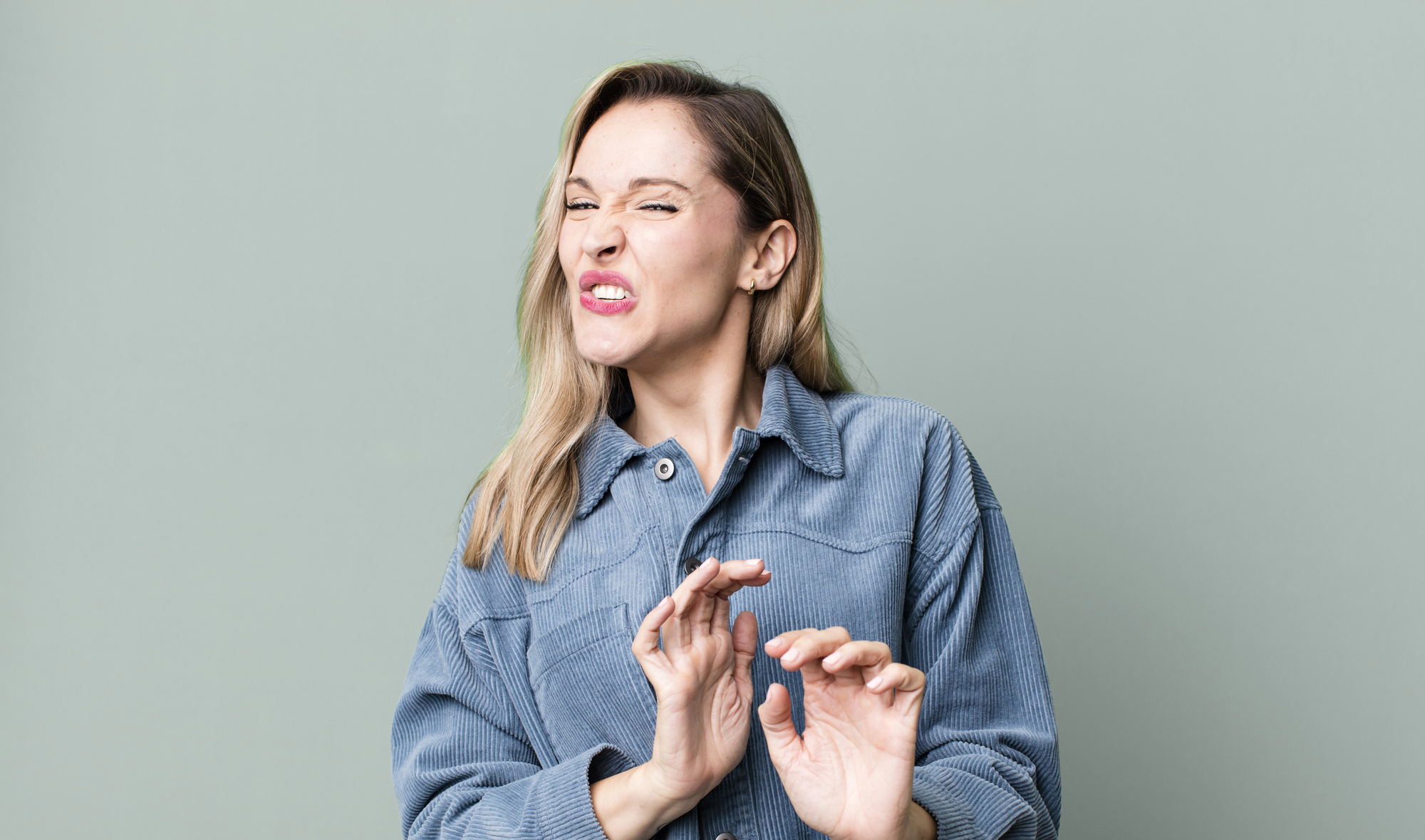 A person with long hair and a denim shirt makes a disgusted facial expression, with their hands raised in front of them, against a plain light green background.