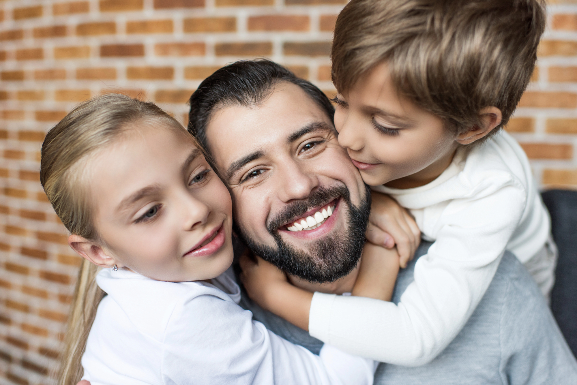 A man with a beard smiles warmly as two children hug him from each side. The group is seated in front of a brick wall, wearing casual clothing. The children appear joyful, showcasing a happy, close-knit family moment.