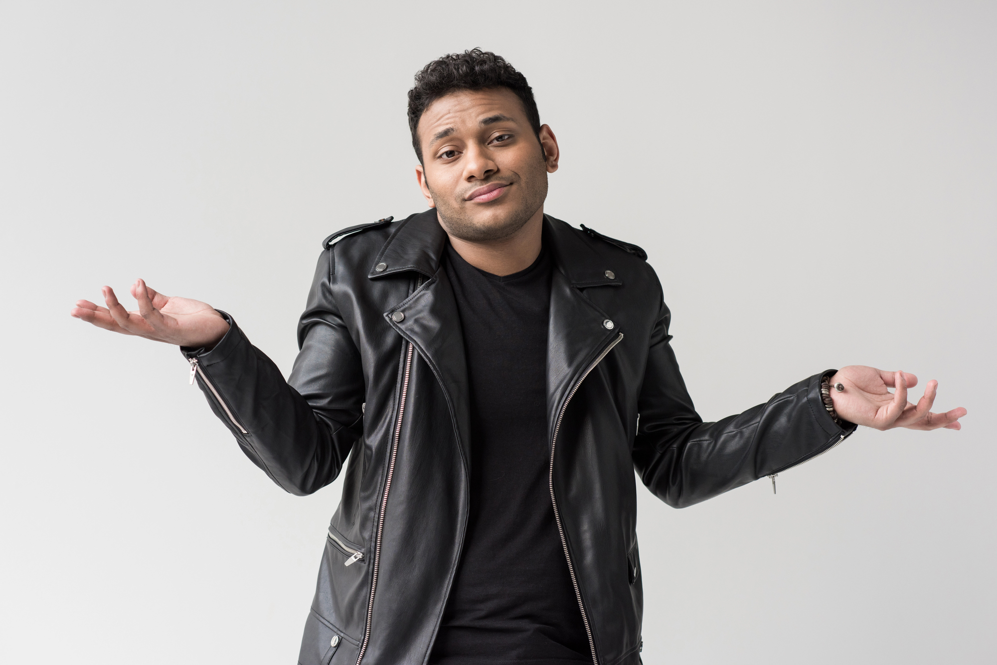 A person wearing a black leather jacket and a black shirt stands against a plain background, shrugging with both hands raised and a slight smile, conveying a sense of uncertainty or indifference.